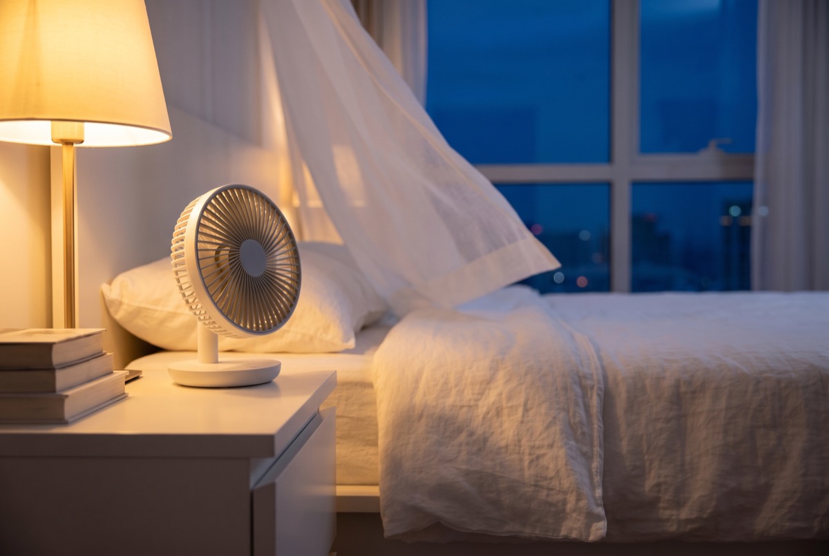 Kleine ventilator op een nachtkastje die over het bed blaast en het laken optilt, warm lamplicht en nacht buiten