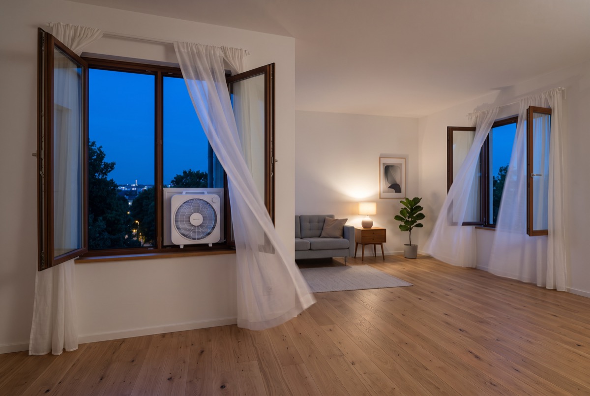 Raamventilator in een openstaand raam bij avondlicht, met aan de andere kant van de kamer een tweede raam open